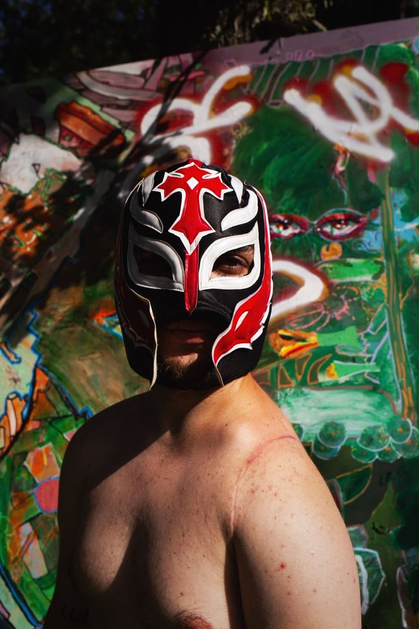 Sage in a black and red wrestling mask in front of a painted wall in Braga